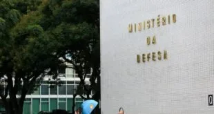 Fachada de prédio com letreiro dourado 'Ministério da Defesa' e pedestre passando em frente