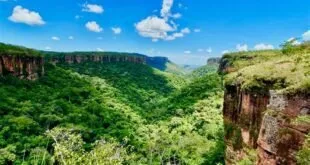 Vale coberto de mata verde com paredão rochoso ao fundo e céu azul com nuvens