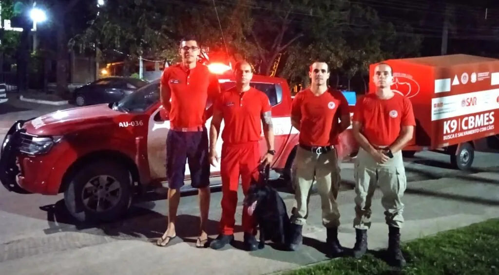 Quatro bombeiros de uniforme vermelho posam à noite com cão ao lado de viatura K9