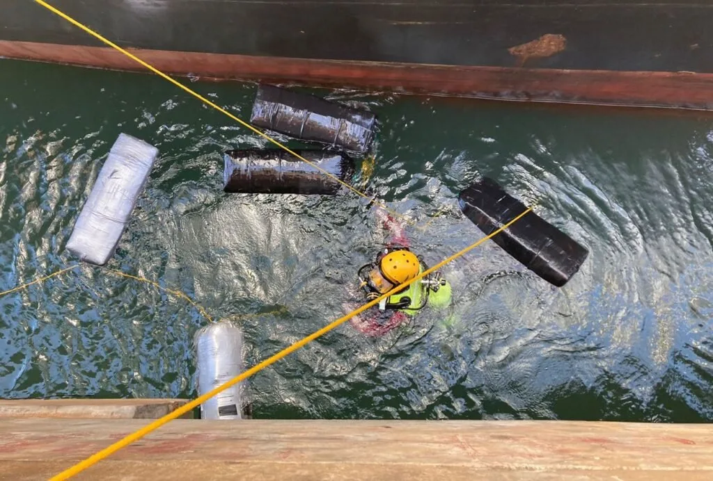 Mergulhador de colete amarelo na água ao lado de fardos escuros presos ao casco de navio