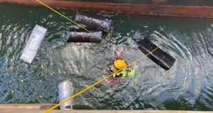 Mergulhador de colete amarelo na água ao lado de fardos escuros presos ao casco de navio