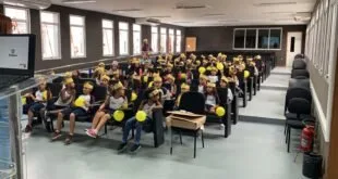 Crianças uniformizadas com chapéus amarelos de papel e balões sentadas em auditório