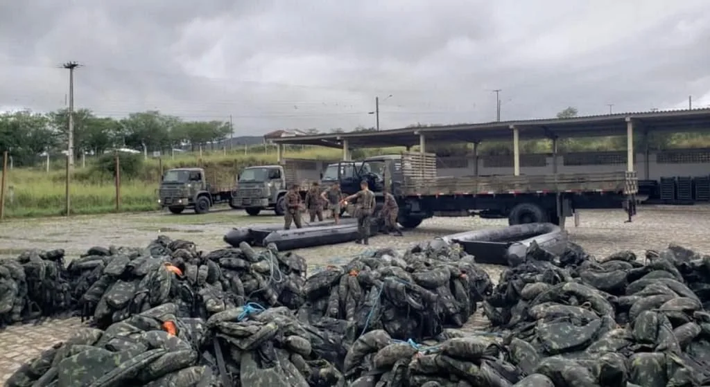 Militares ao lado de caminhões do Exército e pilhas de sacos escuros empilhados no chão