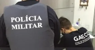 Policial de costas com colete escrito 'Polícia Militar' ao lado de homem de máscara