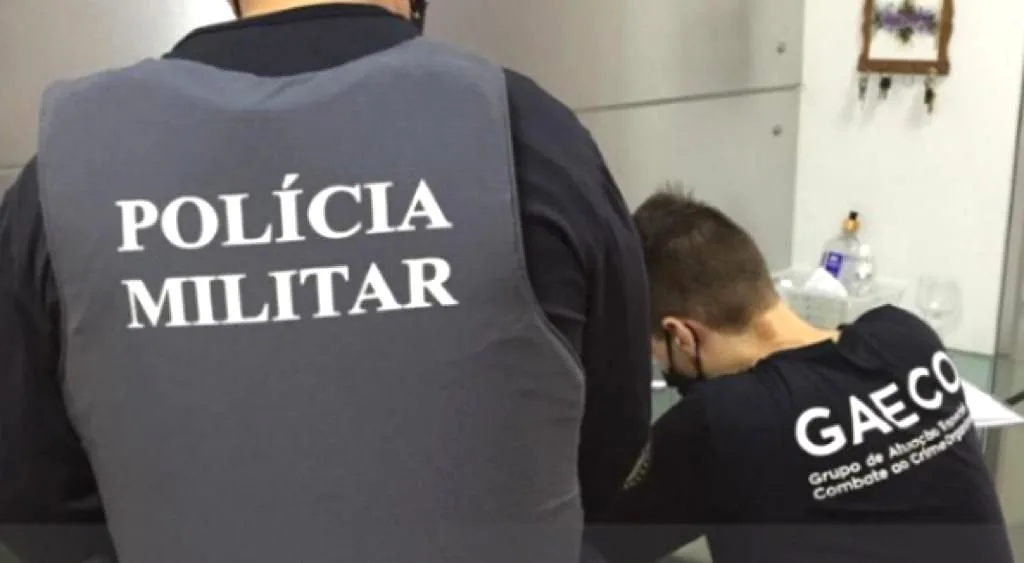 Policial de costas com colete escrito 'Polícia Militar' ao lado de homem de máscara