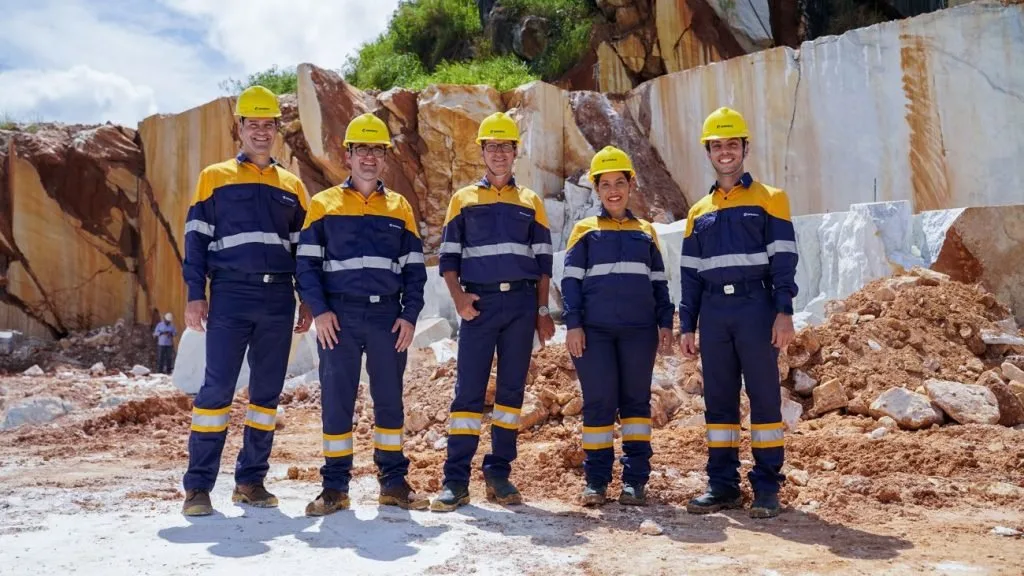 Grupo de trabalhadores de uniforme e capacete amarelo posa diante de pedreira