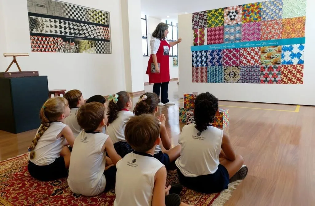 Crianças uniformizadas sentadas no chão de galeria ouvem apresentação diante de obras coloridas
