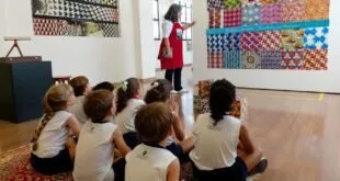 Crianças uniformizadas sentadas no chão de galeria ouvem apresentação diante de obras coloridas