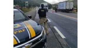 Agente ao lado de viatura da Polícia Rodoviária Federal na margem de rodovia com caminhão ao fundo