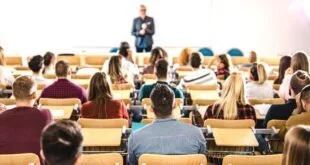 Estudantes sentados em auditório vistos de costas, com professor ao fundo da sala