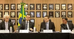 Dois homens de terno à mesa de uma coletiva, com bandeira do Brasil e quadros ao fundo