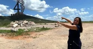 Mulher de preto apontando para uma escultura em terreno com entulho sob céu azul