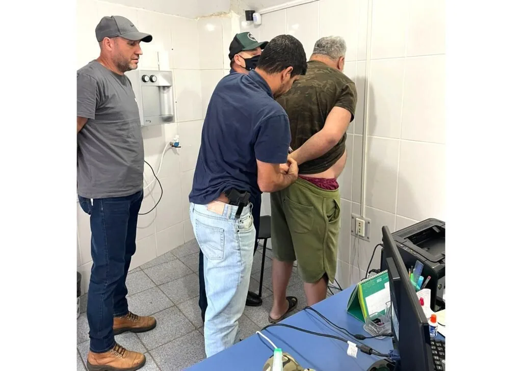 Policiais algemando um homem em sala de azulejos brancos de delegacia