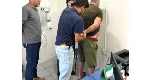 Policiais algemando um homem em sala de azulejos brancos de delegacia