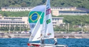 Barco a vela com a bandeira do Brasil na vela navegando com dois velejadores a bordo