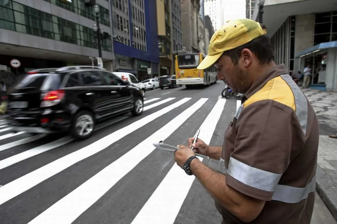 Agente de trânsito de boné amarelo anotando em bloco ao lado de faixa de pedestres