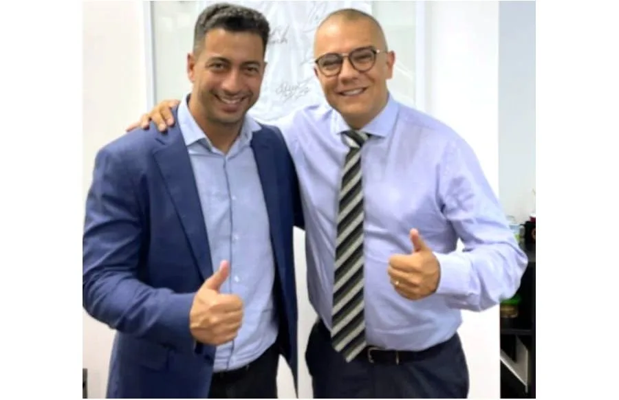 Dois homens de camisa social lado a lado, sorrindo e fazendo sinal de positivo