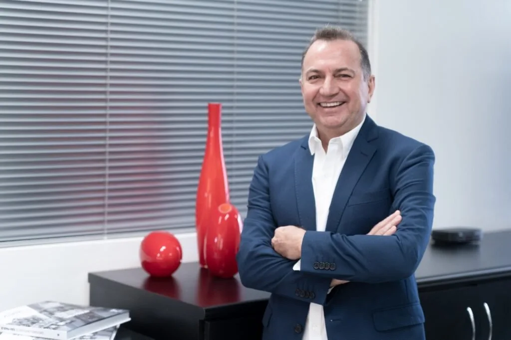 Homem de terno azul sorrindo de braços cruzados ao lado de vasos vermelhos em sala clara