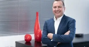 Homem de terno azul sorrindo de braços cruzados ao lado de vasos vermelhos em sala clara