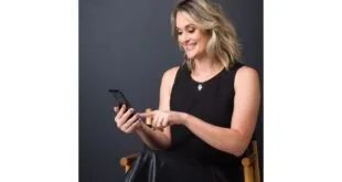 Mulher loira de vestido preto sentada e sorrindo enquanto usa o celular em estúdio