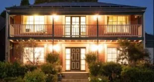 Casa de dois andares com luzes acesas e varanda ao anoitecer, cercada por jardim