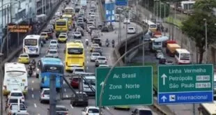 Congestionamento de carros e ônibus em avenida com placas verdes de Zona Norte e Zona Oeste