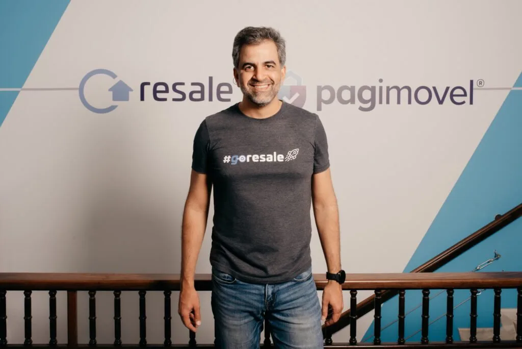 Homem de camiseta escura sorrindo diante de parede com os logotipos 'resale' e 'pagimovel'