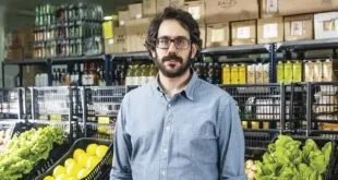 Homem de óculos e camisa azul em pé entre prateleiras de hortifrúti e conservas