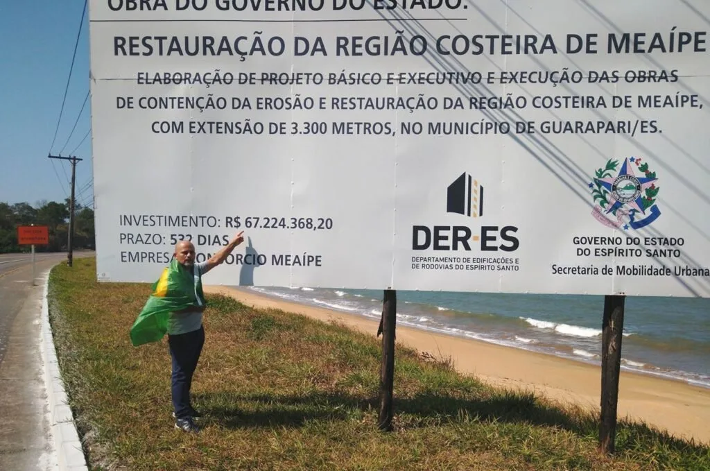 Homem diante de placa verde de obra de restauração da região costeira em área de areia