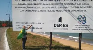 Homem diante de placa verde de obra de restauração da região costeira em área de areia
