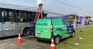 Trabalhador em escada instala equipamento em poste ao lado de van verde e ônibus urbano