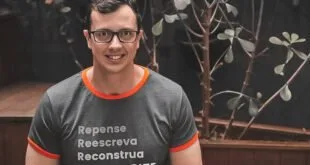 Homem de óculos com camiseta escrita 'Repense, reescreva, reconstrua' em ambiente interno