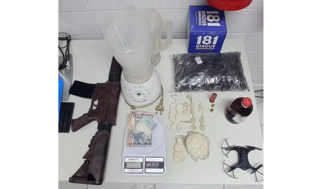Fuzil, drone, porções de droga, balança e cédulas apreendidos sobre mesa branca