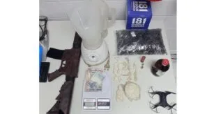 Fuzil, drone, porções de droga, balança e cédulas apreendidos sobre mesa branca