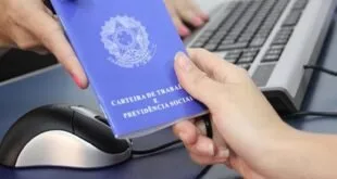 Mãos segurando Carteira de Trabalho e Previdência Social azul sobre mesa com notebook