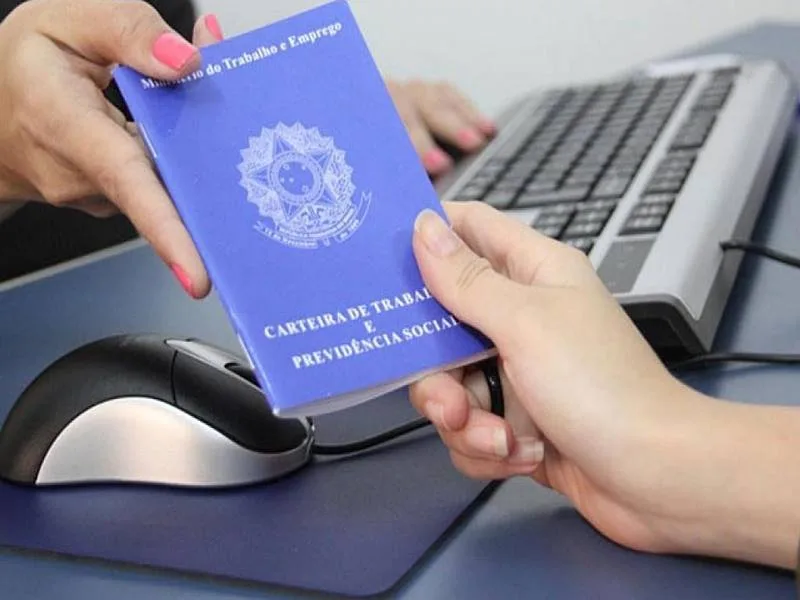 Mãos segurando Carteira de Trabalho e Previdência Social azul sobre mesa com notebook