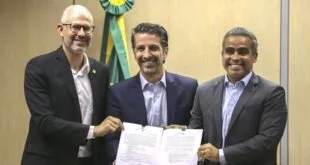 Três homens de terno exibem documento assinado diante de painel com a bandeira do Brasil