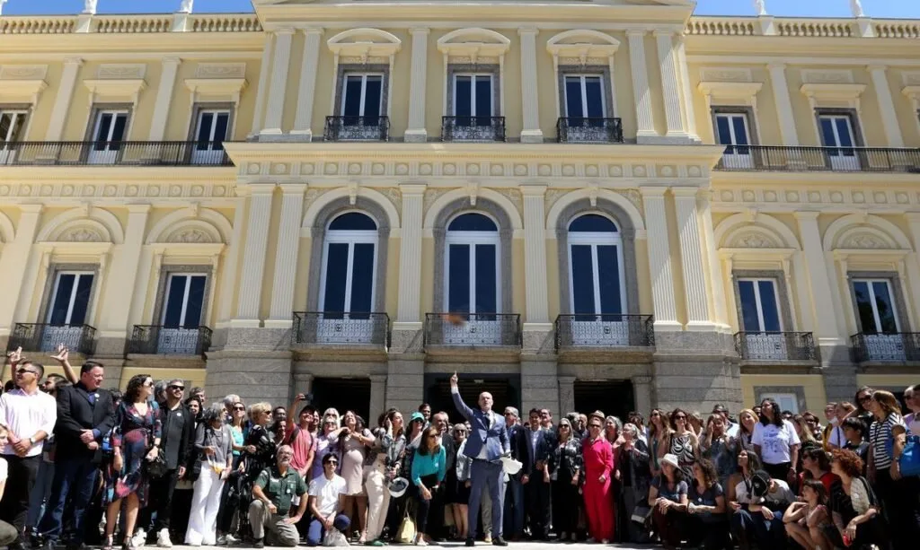 Grupo de pessoas posando na escadaria de palácio amarelo de arquitetura neoclássica