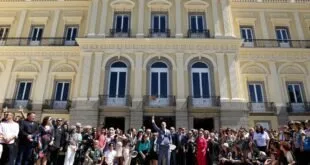 Grupo de pessoas posando na escadaria de palácio amarelo de arquitetura neoclássica