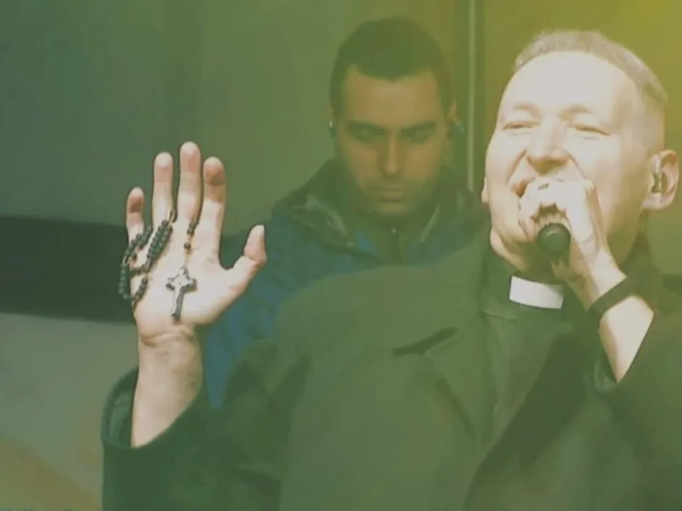 Homem com colarinho clerical segura terço e canta ao microfone