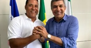 Dois homens sorrindo de mãos dadas diante de bandeiras, entre elas a do Brasil
