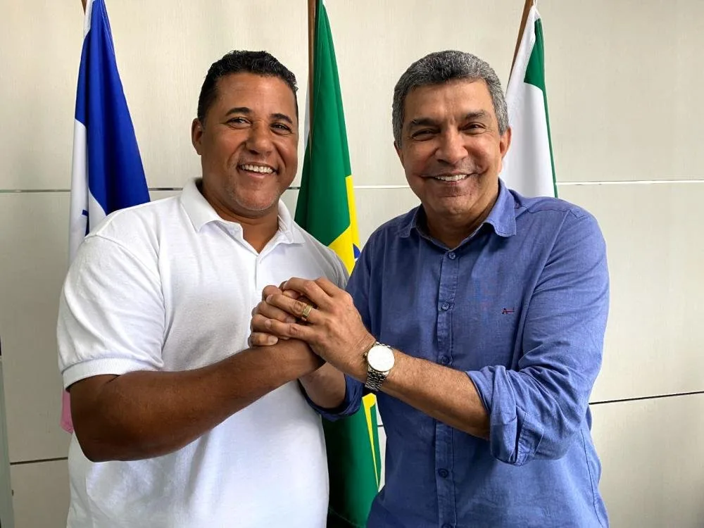Dois homens sorrindo de mãos dadas diante de bandeiras, entre elas a do Brasil
