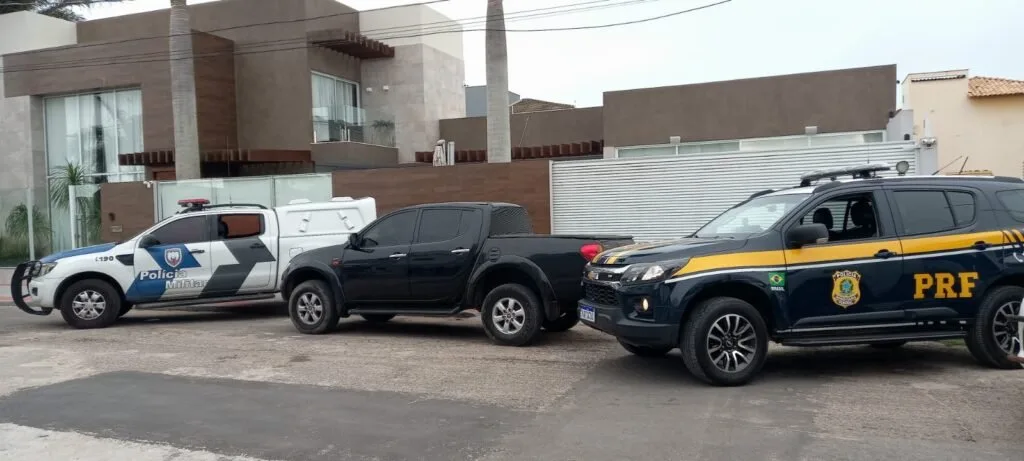 Viaturas da Polícia Militar e da PRF ao lado de caminhonete preta em frente a casa