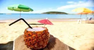 Bebida servida dentro de abacaxi em mesa de praia com guarda-sol e mar ao fundo