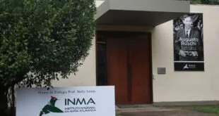Placa com a sigla INMA na entrada de um prédio cercado por árvores
