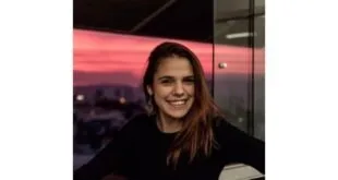 Mulher sorrindo em ambiente escuro diante de janela com céu alaranjado ao entardecer