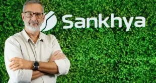 Homem de braços cruzados diante de parede de folhagem com o logotipo Sankhya