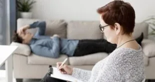 Mulher de óculos anota em caderno enquanto pessoa está deitada em sofá ao fundo