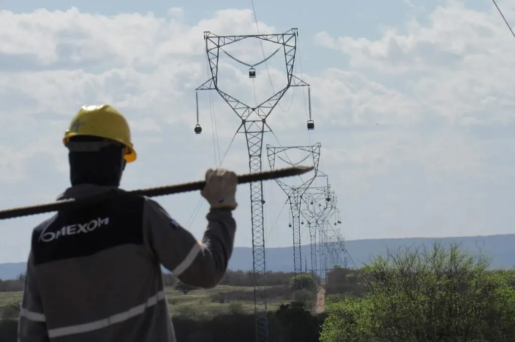 Trabalhador de capacete amarelo carrega cabo diante de torres de linha de transmissão