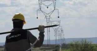 Trabalhador de capacete amarelo carrega cabo diante de torres de linha de transmissão
